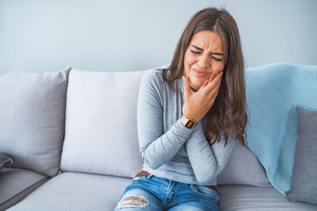 Woman in grey shirt on grey couch pressing hands to jaw in pain